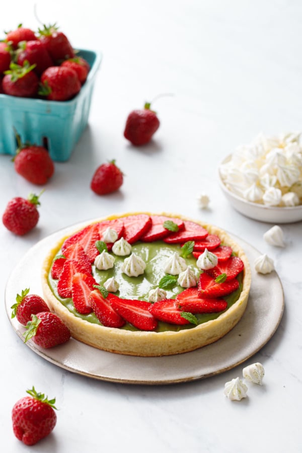 Matcha Strawberry Tartlet on a marble background, with turquoise basket of strawberries and a bowl of mini meringue kisses.