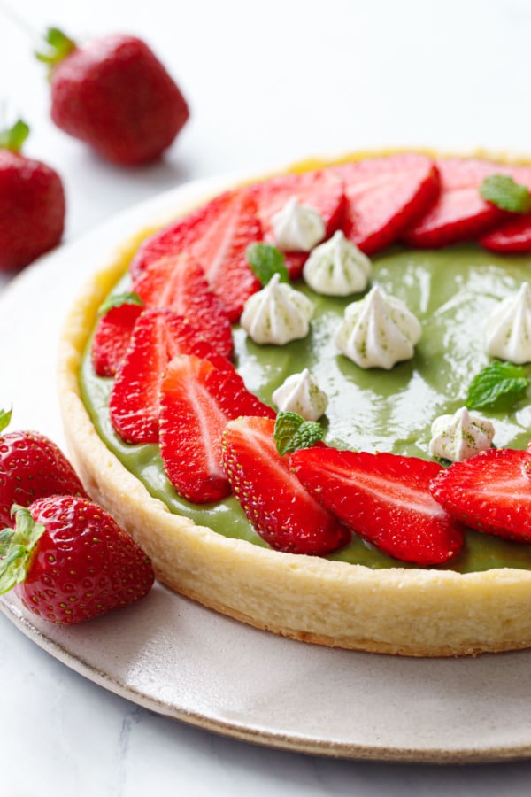 Closeup of green matcha cream filled tart with slices of strawberries arranged in a ring on top, with tiny meringue kisses and fresh strawberries scattered around.