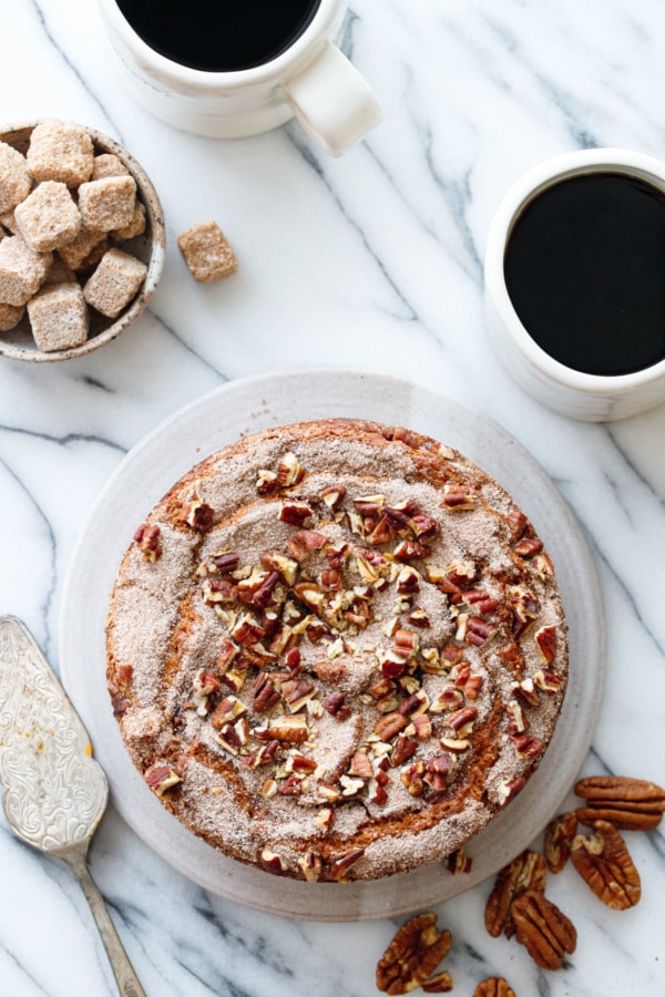 Overhead scene with coffee cake and scattered pecans, two coffee cups and a bowl of sugar cubes