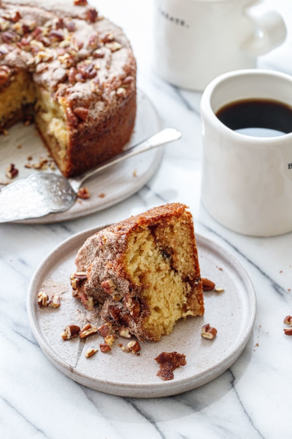 Slice of Greek Yogurt Coffee Cake laying on its side on a plate, with the full cake and coffee cups in the background.