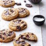 Olive Oil Chocolate Chip Cookies on parchment, one cookie broken in half with a small bowl of chocolate chunks and flaky sea salt