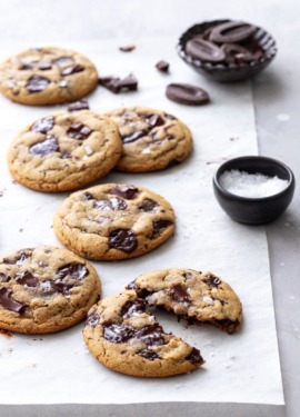 Olive Oil Chocolate Chip Cookies on parchment, one cookie broken in half with a small bowl of chocolate chunks and flaky sea salt