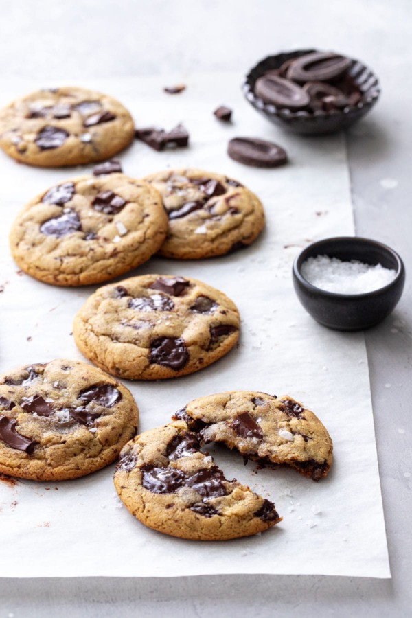 Biscuits aux pépites de chocolat à l'huile d'olive sur papier parchemin, un biscuit cassé en deux avec un petit bol de morceaux de chocolat et de sel de mer feuilleté