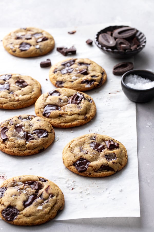 Biscuits aux pépites de chocolat à l'huile d'olive sur parchemin, avec un petit bol de morceaux de chocolat et du sel marin en paillettes