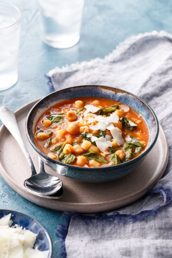 Ceramic bowl with pasta and chickpea stew on a ceramic plate with two spoons and a napkin.