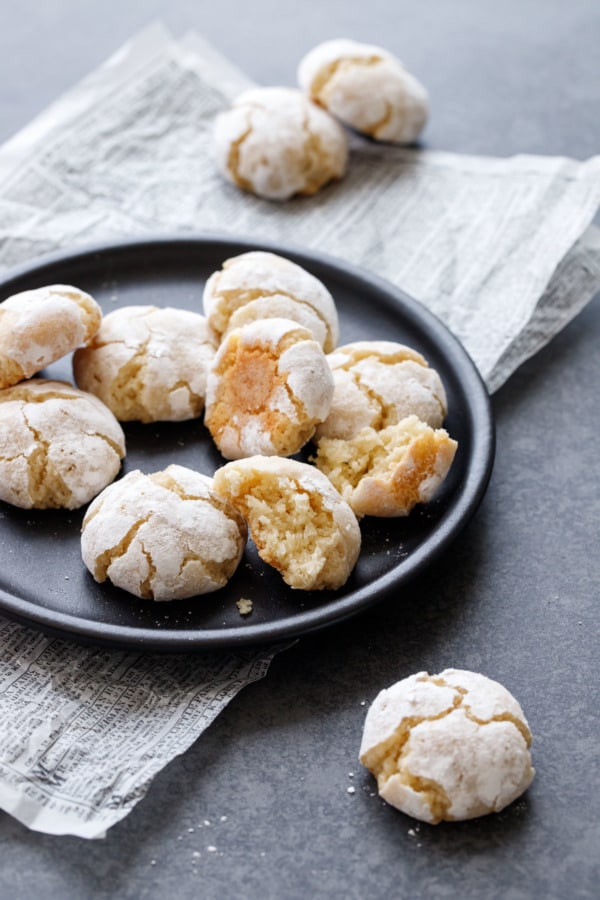 Black plate with amaretti cookies, one broken in half to show texture, on a dark background with newsprint parchment