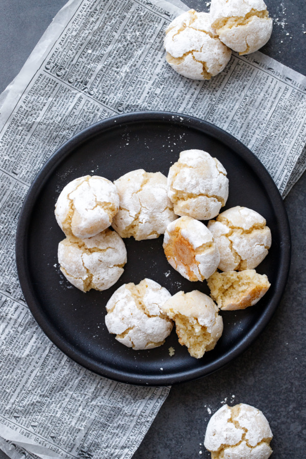 Overhead of a black plate with amaretti cookies, one broken in half to show the chewy texture.