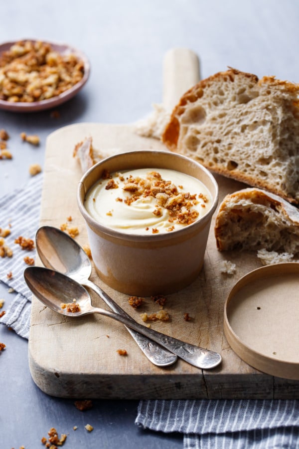 Half pint container of Sourdough Ice Cream topped with breadcrumbs, on a wooden board with spoons and torn up pieces of sourdough bread.