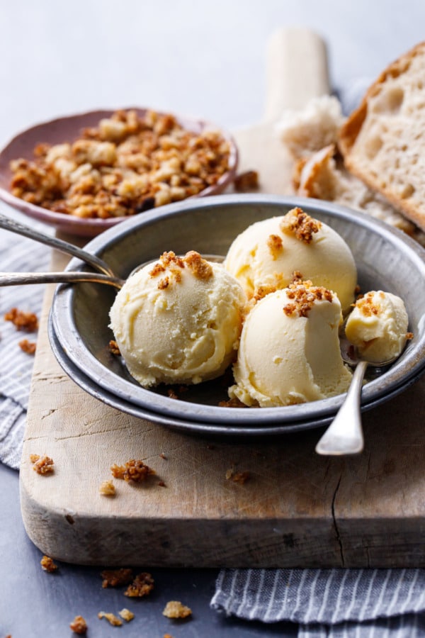 Three scoops of Sourdough Ice Cream in a metal pie tin, on a wood bread board with chunks of sourdough bread and a dish of breadcrumbs.