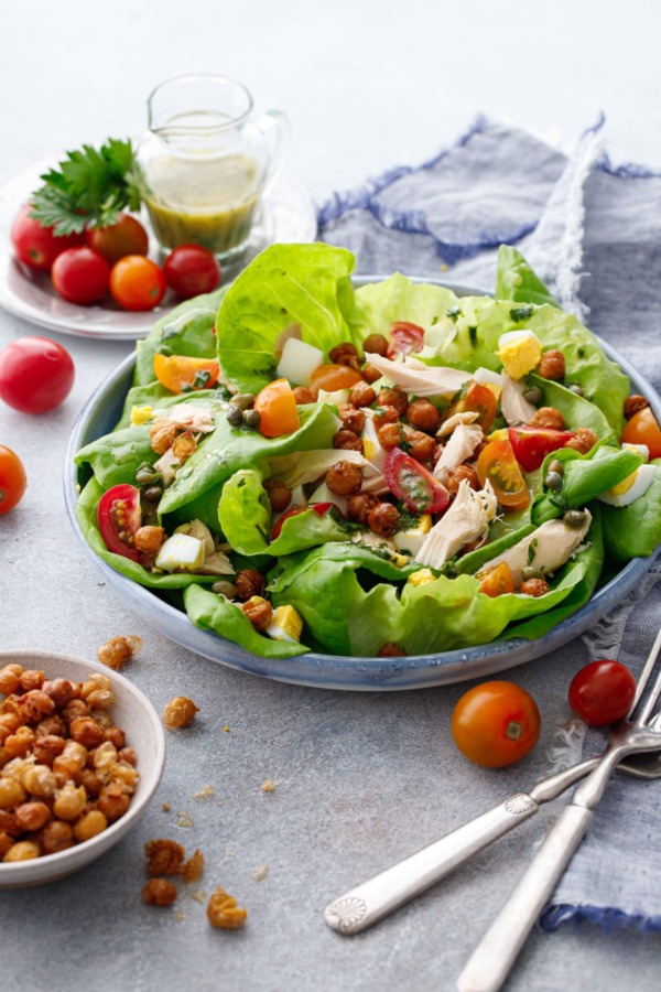 Large shallow dish with butter lettuce and nicoise salad toppings, with a dish of crispy chickpeas and tomatoes and dressing in the background.