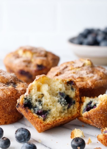 Split blueberry muffin to show inside texture, on marble with more muffins and blueberries in the background