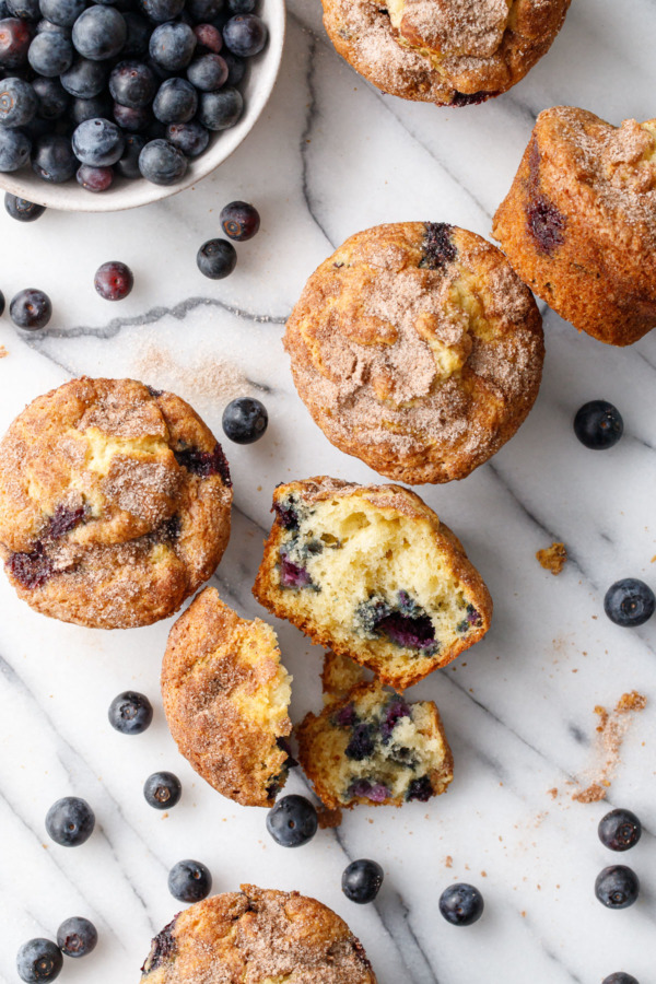 Overhead of scattered muffins and blueberries on marble, one muffin split in half to show the fluffy texture.