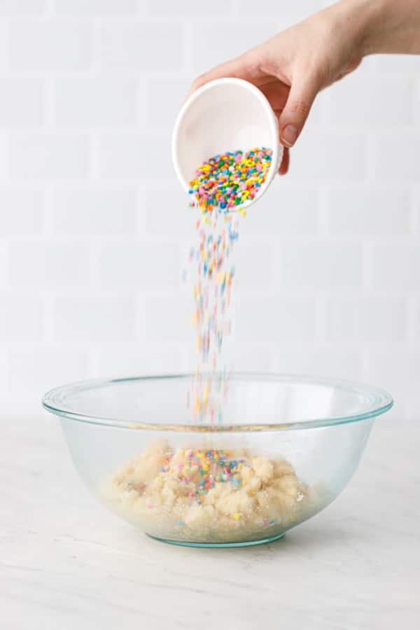 Pouring rainbow sprinkles into a glass bowl of amaretti cookie dough