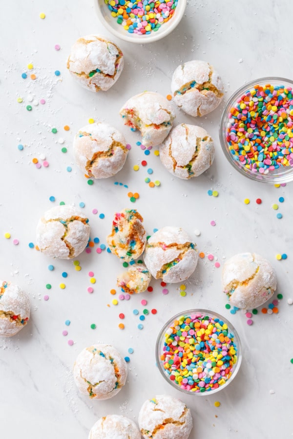 Overhead of scattered amaretti cookies, three bowls of sprinkles plus more scattered around