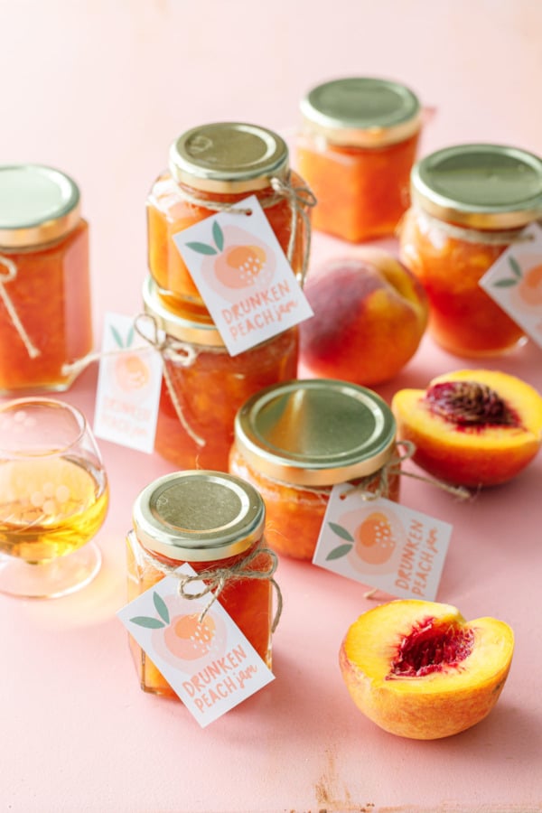 Glass jars of orange peach jam on a pink background, with a small glass of bourbon and peaches cut in half.