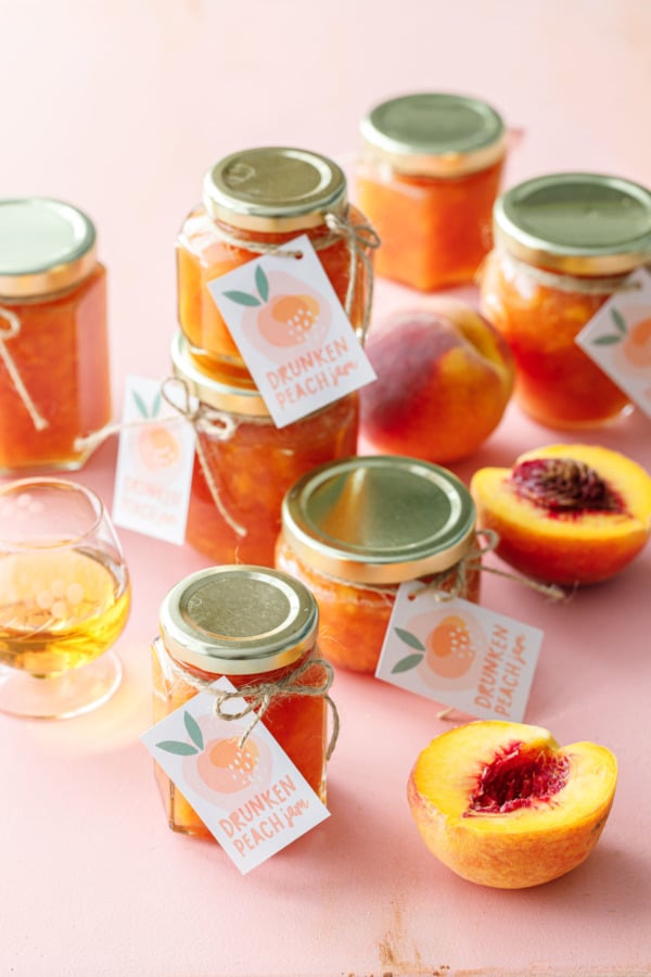 Bocaux en verre de confiture de pêche orange sur fond rose, avec un petit verre de bourbon et des pêches coupées en deux.