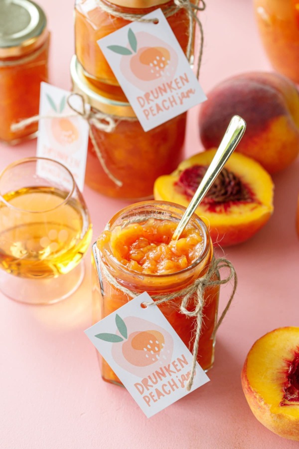Open jar of peach jam with a gold spoon, on a pink background with more jars of jam, cut peaches and a glass of bourbon.