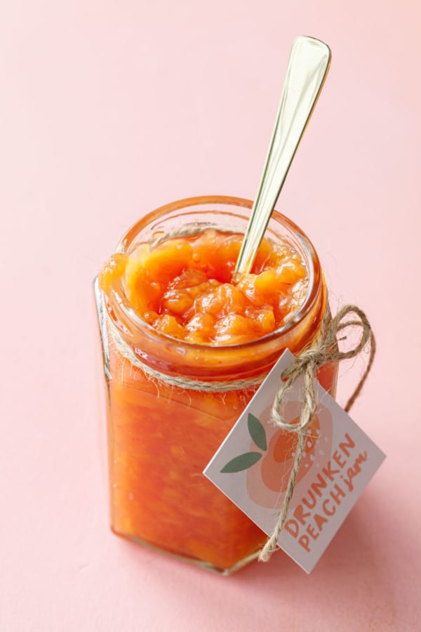 Open glass jar of drunken peach jam with a gold spoon and hang tag label, showing the perfect texture of the jam.