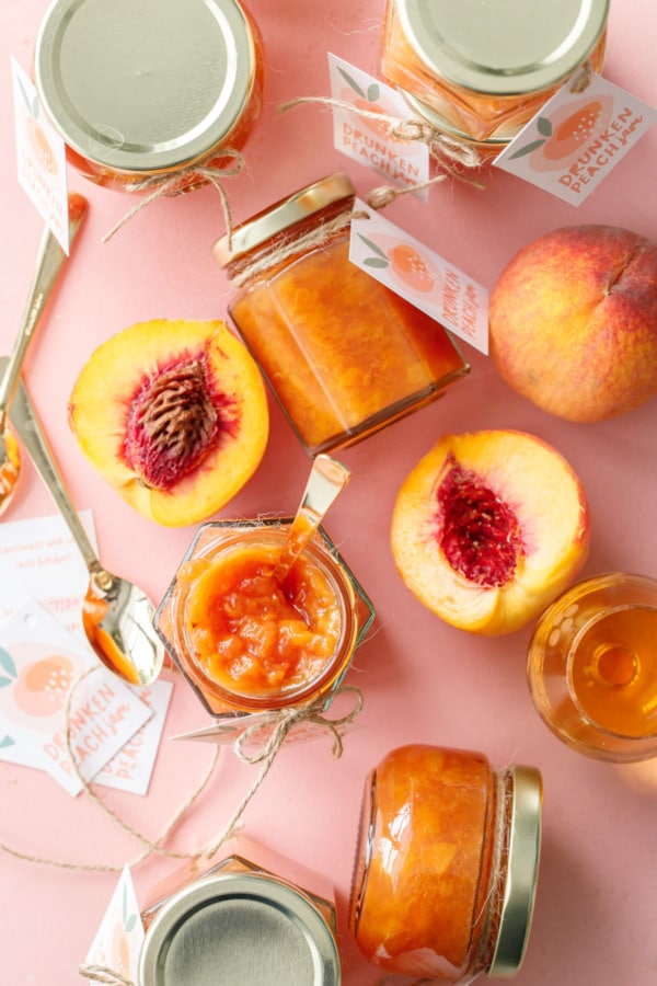 Overhead of various jars of drunken peach jam, one jar open, with cut peaches, a glass of bourbon, and printed tags scattered around.