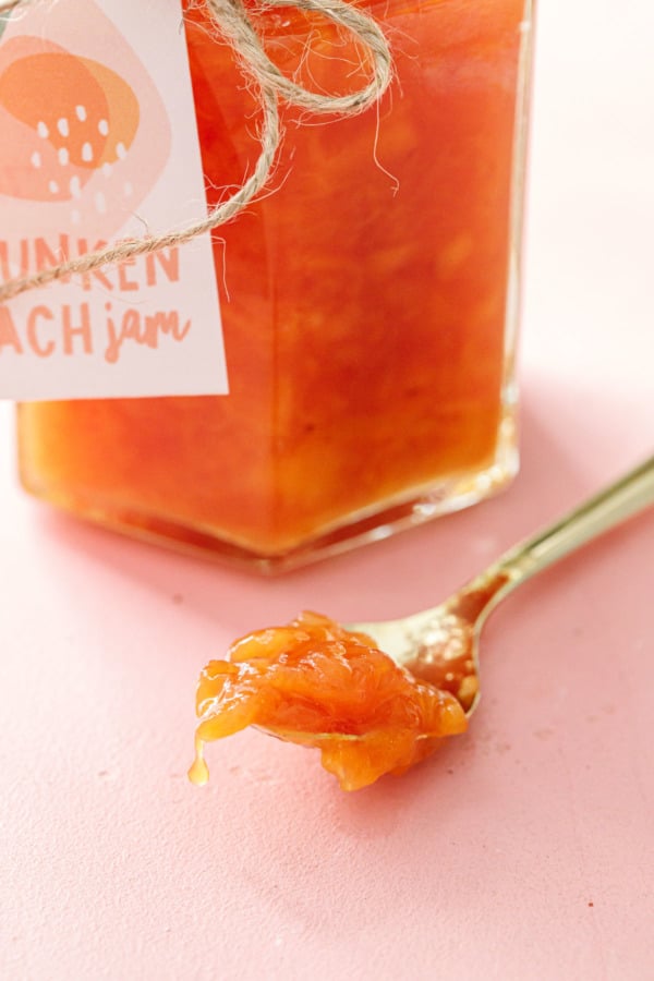 Closeup of a spoonful of drunken peach jam to show the texture, on a pink background with a jar of jam in the back.