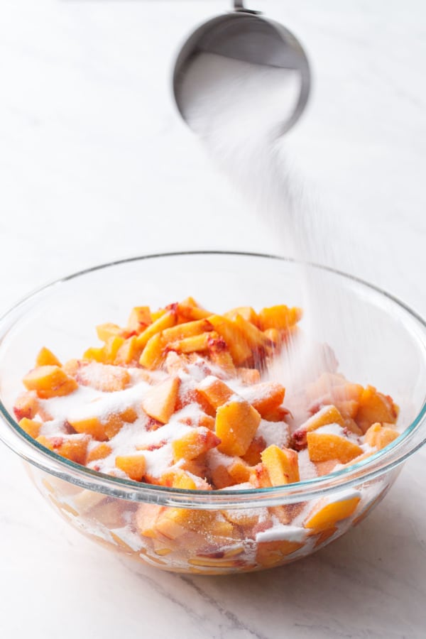 Pouring sugar over chopped peaches in a glass bowl