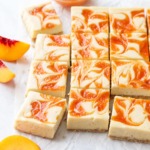 Peach cheescake bars cut into squares, arranged on a piece of crinkled parchment with a bowl of peach puree and a few peach slices scattered around.