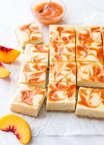 Peach cheescake bars cut into squares, arranged on a piece of crinkled parchment with a bowl of peach puree and a few peach slices scattered around.