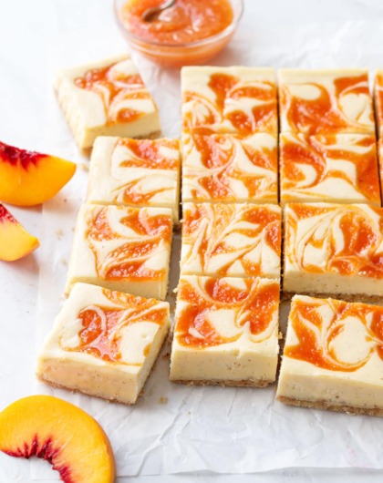 Peach cheescake bars cut into squares, arranged on a piece of crinkled parchment with a bowl of peach puree and a few peach slices scattered around.