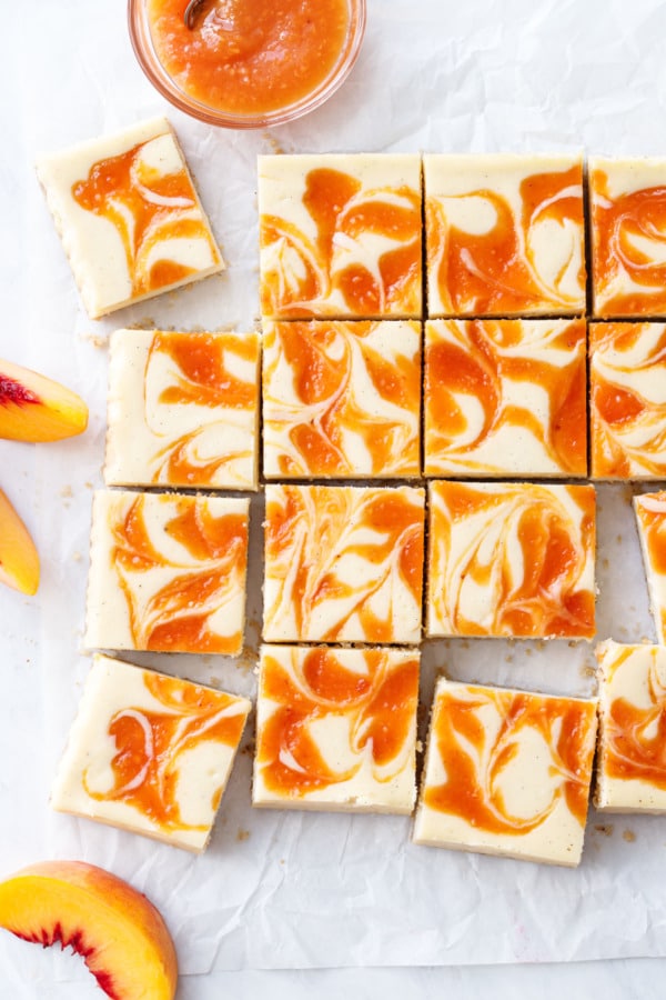 Overhead of peach cheesecake bars cut into squares with swirled peach puree baked on top; on crinkled parchment paper with a bowl of peach puree and peach slices.