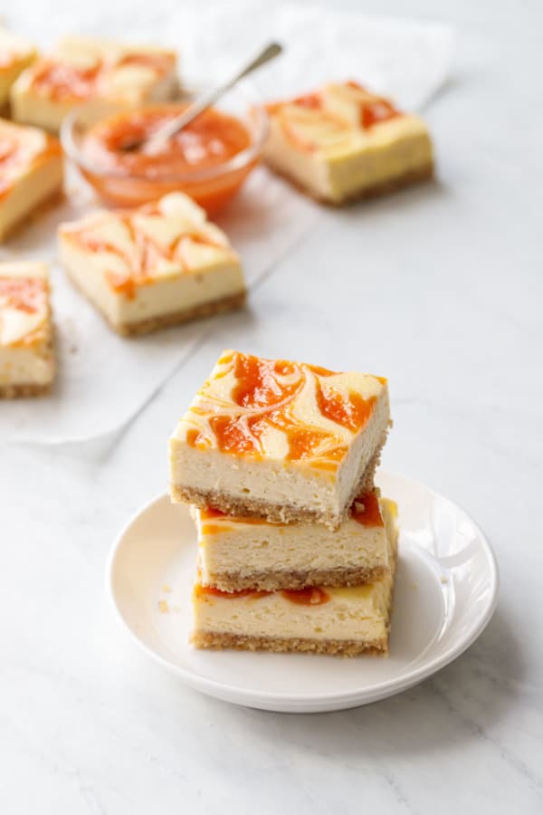 Stack of 3 peach cheesecake bars on a small white plate, with more cut bars and a bowl of peach puree in the background