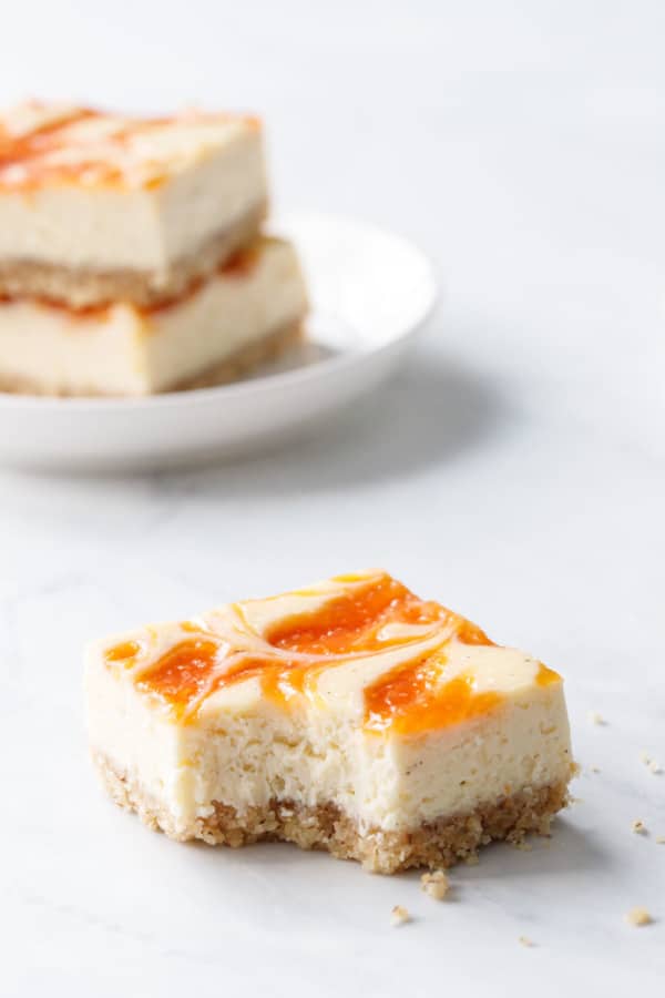 One square of peach cheesecake with a bite taken out of it, on a marble background with a stack of two more bars in the background.