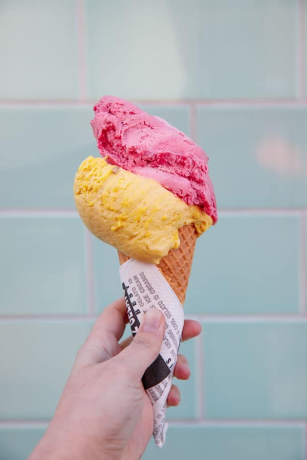 Cone with scoops of mango and cherry gelato against a blue tile background