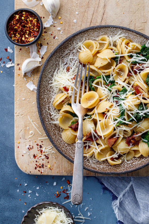 Overhead, bowl of pasta with bacon and wilted frisee on a wooden board with pepper flakes and pecorino cheese