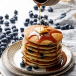 Drizzling maple syrup on a tall stack of blueberry pancakes