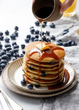 Drizzling maple syrup on a tall stack of blueberry pancakes