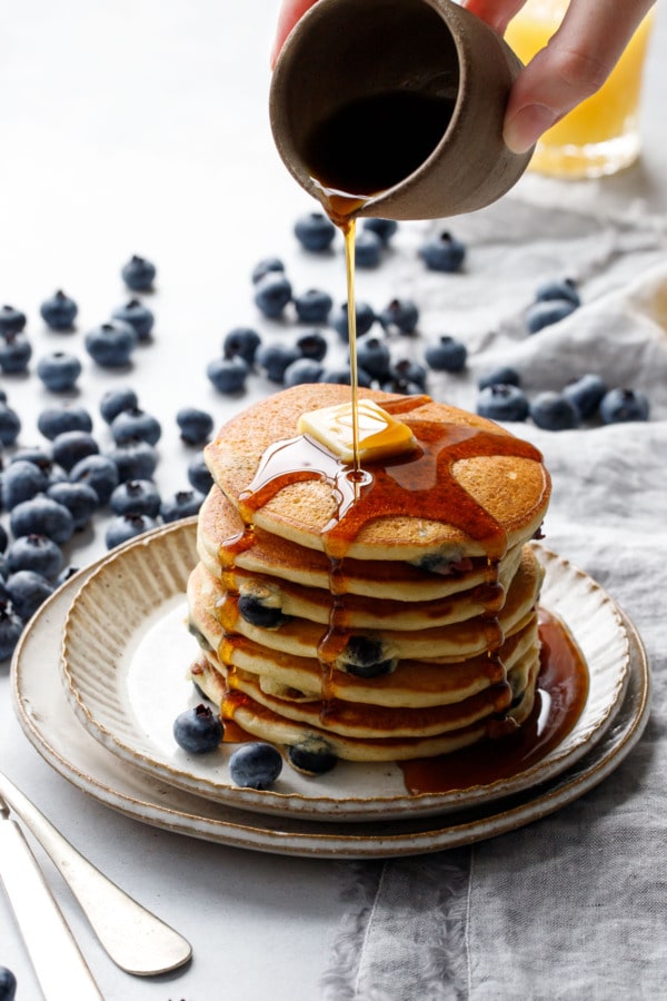 Drizzling maple syrup on a tall stack of blueberry pancakes