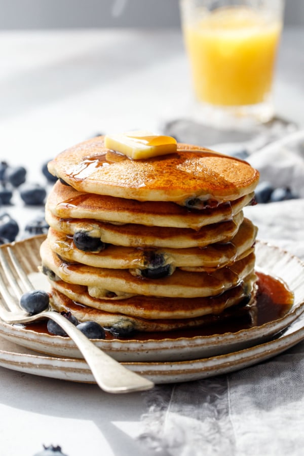 Stack of blueberry pancakes with a pool of syrup on a plate, bright sunlight