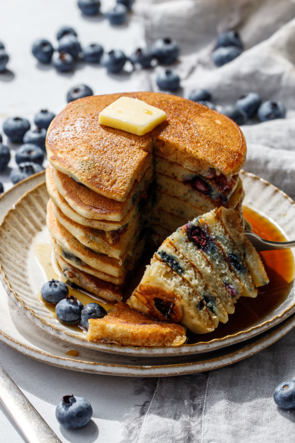 Wedge cut out of a stack of blueberry pancakes, on a plate with a puddle of maple syrup