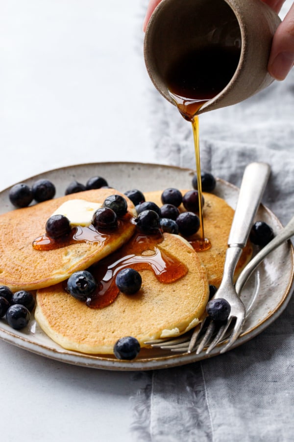 Drizzling maple syrup on a plate of blueberry sourdough pancakes and fresh blueberries