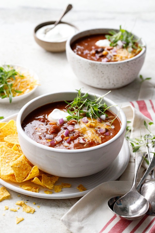 Two white bowls of Vegetarian Pumpkin & Three-Bean Chili topped with cheese, sour cream and micro cilantro