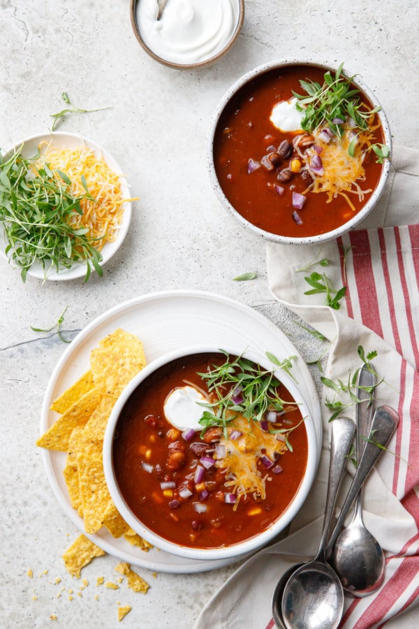 Overhead, two bowls of Vegetarian Pumpkin & Three-Bean Chili with tortilla chips and other toppings