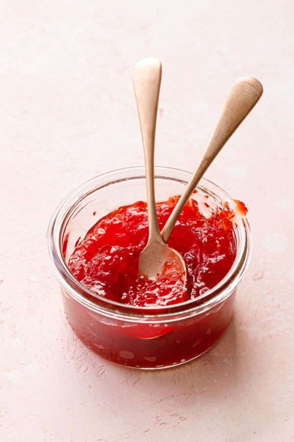 Glass jar of Cranberry Pepper Jelly on a light pink background with two rose-gold spoons