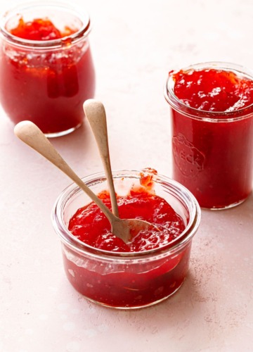 Three glass jars of Cranberry Pepper Jelly on a light pink background, one har with two spoons.