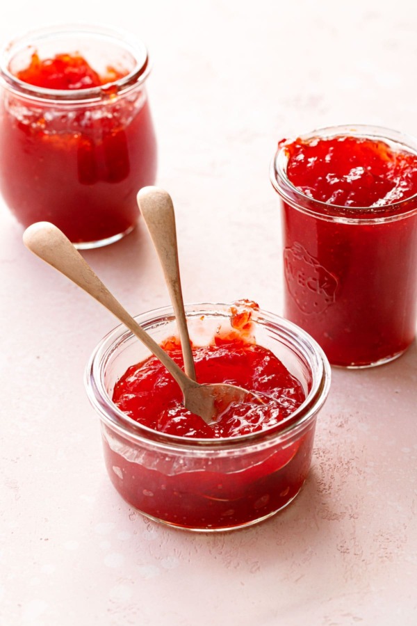 Three glass jars of Cranberry Pepper Jelly on a light pink background, one har with two spoons.