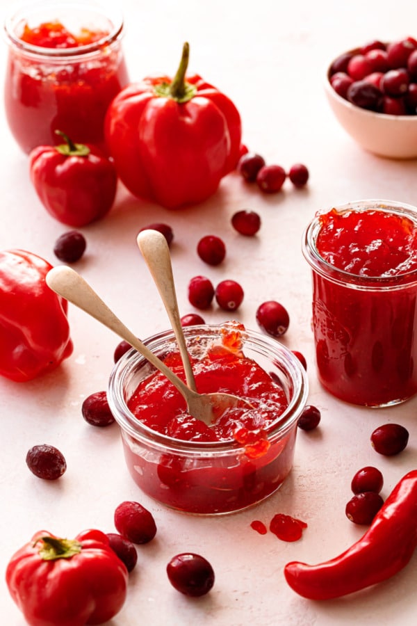 Three glass jars of Cranberry Pepper Jelly on a light pink background, with fresh cranberries and mixed red peppers scattered around.