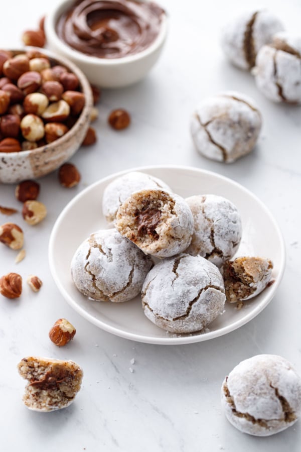 Small plate of Nutella-stuffed Hazelnut Amaretti Cookies, with bowls of hazelnuts and nutella on the side