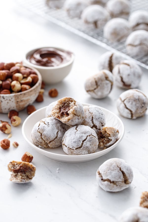 Small plate of Nutella-stuffed Hazelnut Amaretti Cookies, with bowls of hazelnuts and nutella on the side