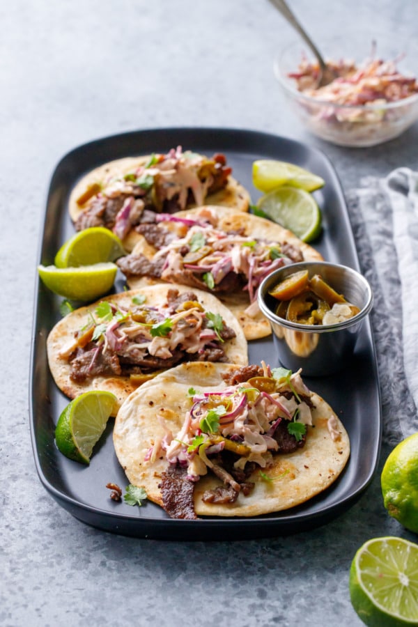 Four steak street tacos on a black rectangular plate, with limes, pickled jalapenos and chipotle slaw