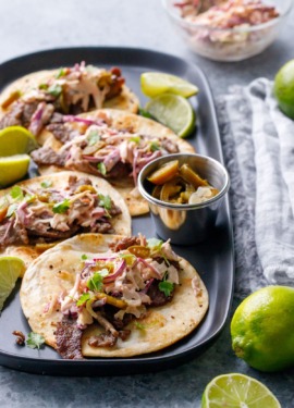 Four flat tacos with steak, slaw and cilantro, slices of limes on the side