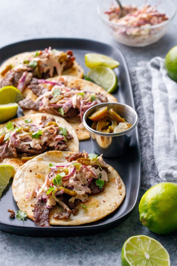 Four flat tacos with steak, slaw and cilantro, slices of limes on the side